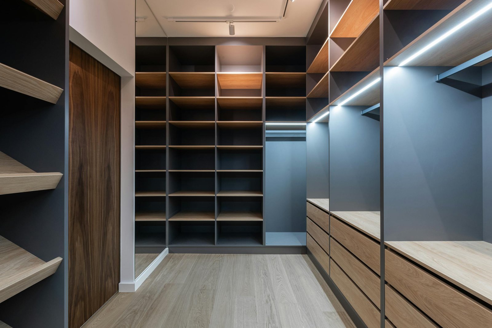 Illustrative image: a walk-in dressing room in oak and grey — open shelving, fitted drawers, integrated lighting
